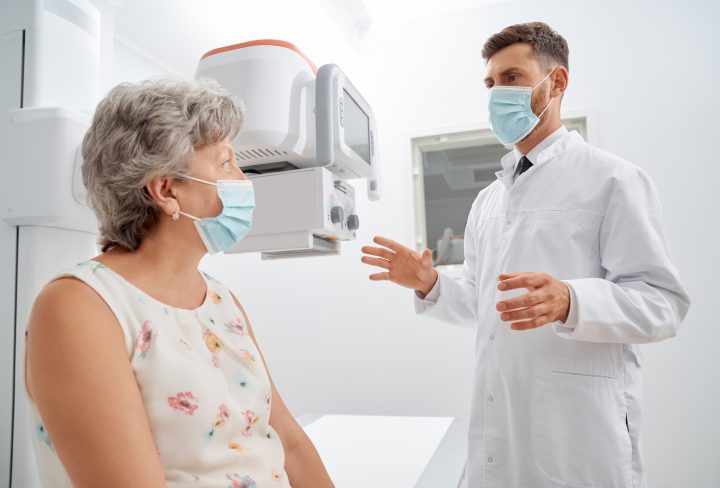 Grey haired woman wearing floral dress on procedure of ultrasound, when radiologist gesturing by hands and preparing. Female patient in cabinet of radiology doctor in uniform telling about process.